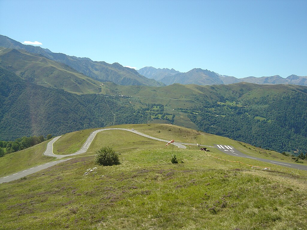 A winding mountain road leads to a small, grassy airfield with a single runway.